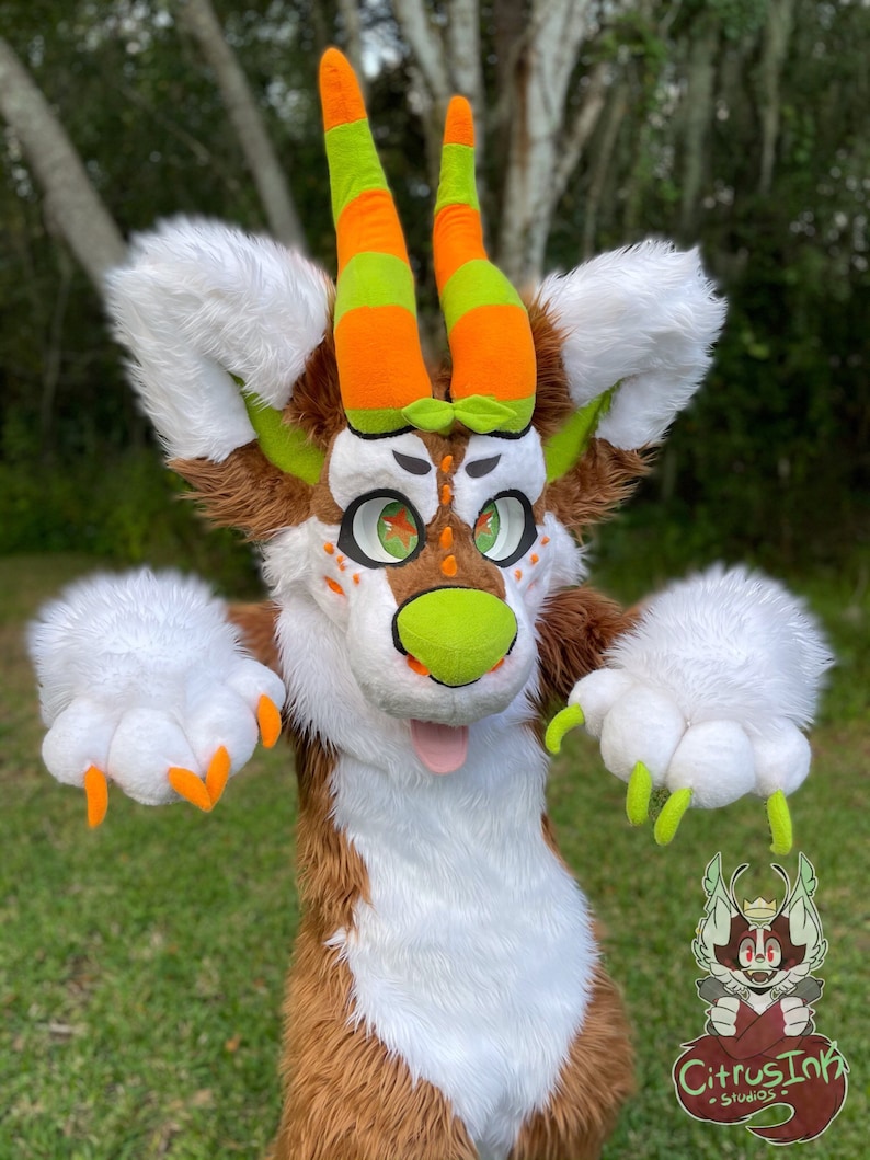 May include: A brown and white furry creature with green and orange horns. The creature has large green eyes and a bright orange tongue sticking out. The creature is wearing a green and orange striped collar. The creature is standing in front of a green grassy background.