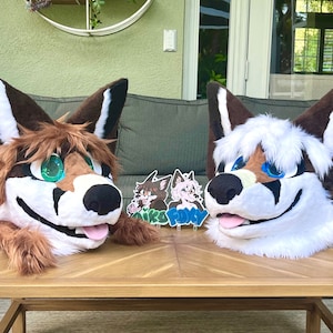 May include: Two furry animal head costumes, one brown and white with green eyes and one white with blue eyes, are sitting on a wooden table.  Two small round stickers with the words "Aiko" and "Foxy" are on the table in front of the costumes.