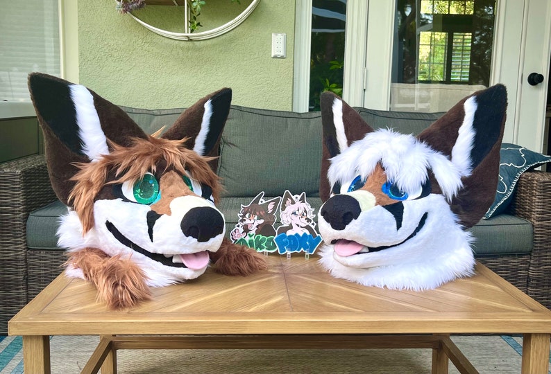 May include: Two furry animal head costumes, one brown and white with green eyes and one white with blue eyes, are sitting on a wooden table.  Two small round stickers with the words "Aiko" and "Foxy" are on the table in front of the costumes.