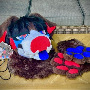 May include: A brown and black furry animal head with a red, white, and blue face. The head has a blue tongue sticking out. The head is on a wooden table with a pair of brown furry paws with red and blue paw pads.