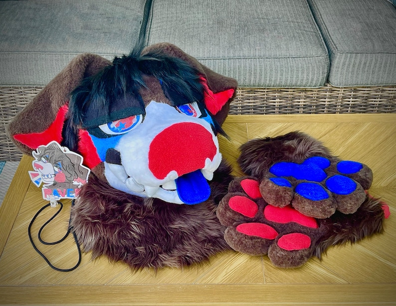 May include: A brown and black furry animal head with a red, white, and blue face. The head has a blue tongue sticking out. The head is on a wooden table with a pair of brown furry paws with red and blue paw pads.