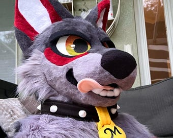 May include: A gray and red wolf fursuit head featuring large pointed ears, yellow eyes with mesh pupils, and a black nose. The character has a pink tongue sticking out, white teeth, and wears a black spiked collar with a yellow tag marked with an M.