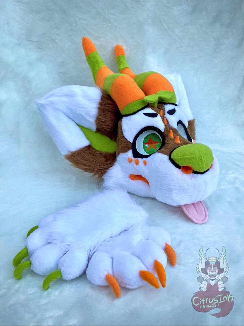 May include: A white, brown, and green furry dragon head with orange and green striped horns. The dragon has a pink tongue sticking out and green eyes with a star-shaped pupil. The dragon head is laying on a white fluffy surface. A white furry paw with green and orange claws is also pictured.