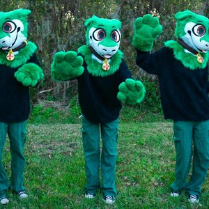 May include: Three green fursuit heads with black sweaters and green pants. The fursuits have green fur and white faces with black eyes and pink noses.