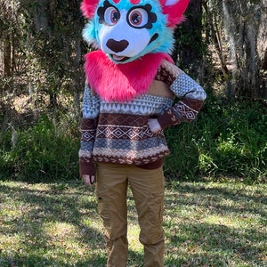 May include: A furry blue and pink animal head with a white face and black nose. The head is wearing a pink fur collar and a brown, white, and grey patterned sweater. The animal is standing in a grassy area.