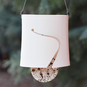 May include: A hanging white ceramic lantern with a curved cutout design and a speckled, circular base. The lantern is suspended by a thin wire and features a small hole near the top. The base has brown speckles.