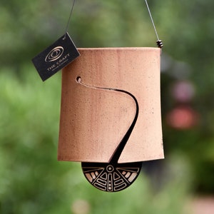 May include: A terracotta-colored wind chime with a black decorative bottom and a curved cutout design. The wind chime is suspended by a wire and includes a tag that reads "THE CRAFT Wind Chimes."