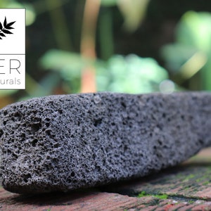 May include: A dark gray, porous lava stone with a rectangular shape. The stone has a rough texture and is set against a blurred green and brown background. The logo for LER Naturals is in the upper left corner.