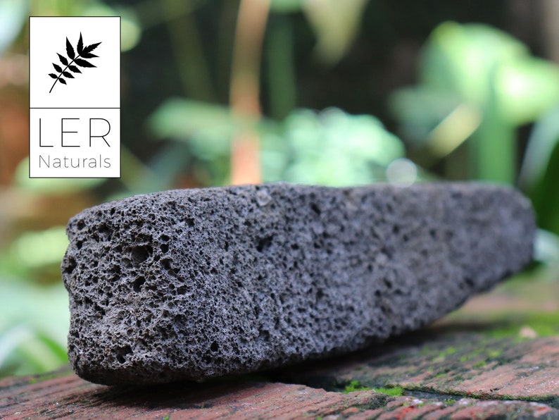 May include: A dark gray, porous lava stone with a rectangular shape. The stone has a rough texture and is set against a blurred green and brown background. The logo for LER Naturals is in the upper left corner.