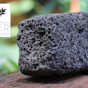 May include: A close-up of a dark gray pumice stone with a porous texture. The stone is rectangular and sits on a reddish-brown surface. The LER Naturals logo is in the upper left corner.