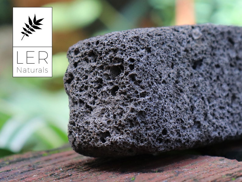 May include: A close-up of a dark gray pumice stone with a porous texture. The stone is rectangular and sits on a reddish-brown surface. The LER Naturals logo is in the upper left corner.