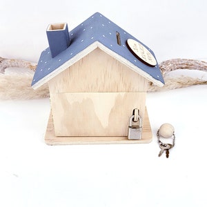 May include: A wooden house-shaped piggy bank with a blue roof and white polka dots. The piggy bank has a slot on the roof and a lock and key on the side. The piggy bank is sitting on a white surface.