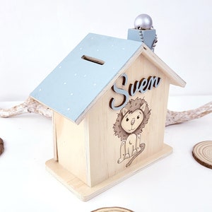 Peut inclure: Tirelire en bois en forme de maison avec un toit bleu clair et des pois blancs. La tirelire a une fente sur le toit pour les pi&egrave;ces de monnaie. Le devant de la tirelire a une illustration de lion et le nom "Sven" &eacute;crit en gris.