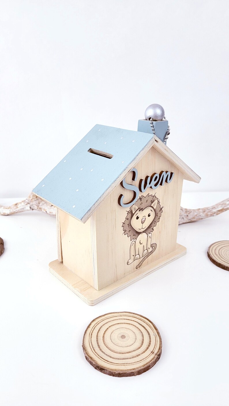 Peut inclure: Tirelire en bois en forme de maison avec un toit bleu clair et des pois blancs. La tirelire a une fente sur le toit pour les pi&egrave;ces de monnaie. Le devant de la tirelire a une illustration de lion et le nom "Sven" &eacute;crit en gris.