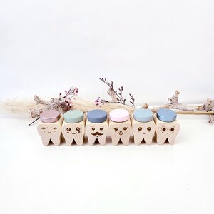 May include: Six wooden teeth with different facial expressions and colored tops. The teeth are arranged in a row on a white surface.