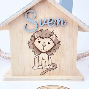 Peut inclure: Une tirelire en bois en forme de maison avec un toit bleu clair et une illustration de lion. Le nom "Sven" est &eacute;crit sur le devant en lettres bleues claires.