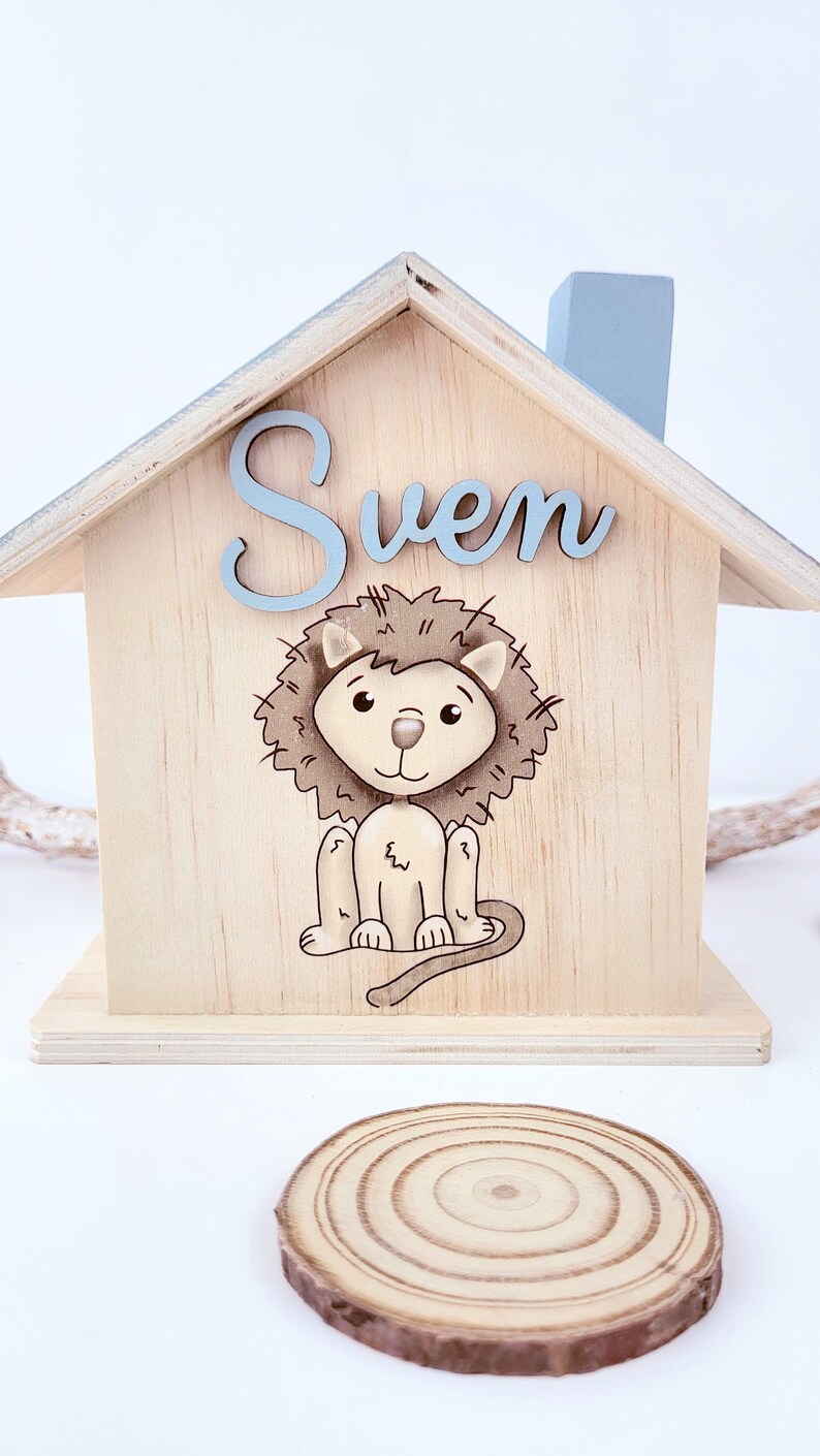 Peut inclure: Une tirelire en bois en forme de maison avec un toit bleu clair et une illustration de lion. Le nom "Sven" est &eacute;crit sur le devant en lettres bleues claires.