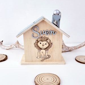 Peut inclure: Une tirelire en bois en forme de maison avec un toit bleu clair et une illustration de lion. Le nom "Sven" est &eacute;crit sur la fa&ccedil;ade de la maison.