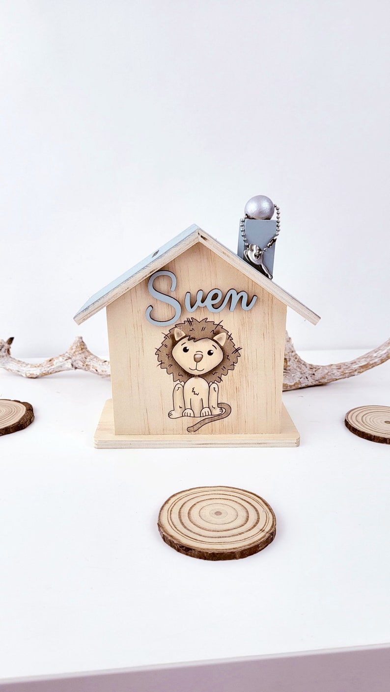 Peut inclure: Une tirelire en bois en forme de maison avec un toit bleu clair et une illustration de lion. Le nom "Sven" est &eacute;crit sur la fa&ccedil;ade de la maison.