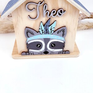 May include: Wooden house shaped piggy bank with a raccoon face peeking out. The house has a gray roof and chimney. The name "Theo" is written on the front of the house.