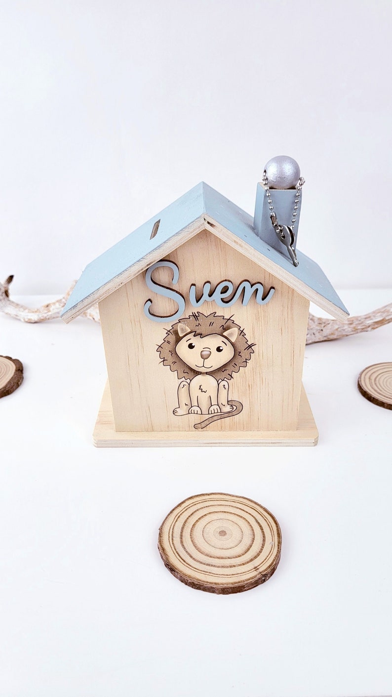 Personalisierte Spardose aus Holz in Form eines Hauses mit hellblauem Dach und Schornstein. Auf der Vorderseite befindet sich ein erhabener 3D-Schriftzug aus Holz mit dem Namen „Sven“ über der Illustration eines niedlichen, sitzenden Löwen mit brauner Mähne. Am Schornstein ist ein Schlüssel an einer Kugelkette befestigt.