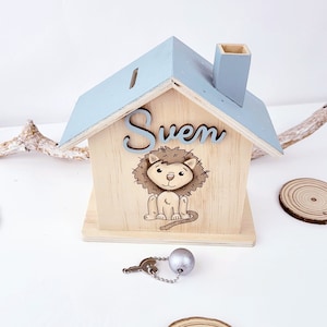 Peut inclure: Une tirelire en bois en forme de maison avec un toit bleu clair et une chemin&eacute;e. Le devant de la tirelire a une illustration de lion et le nom "Sven" &eacute;crit dessus. La tirelire est livr&eacute;e avec une cl&eacute; et une boule argent&eacute;e sur une cha&icirc;ne.