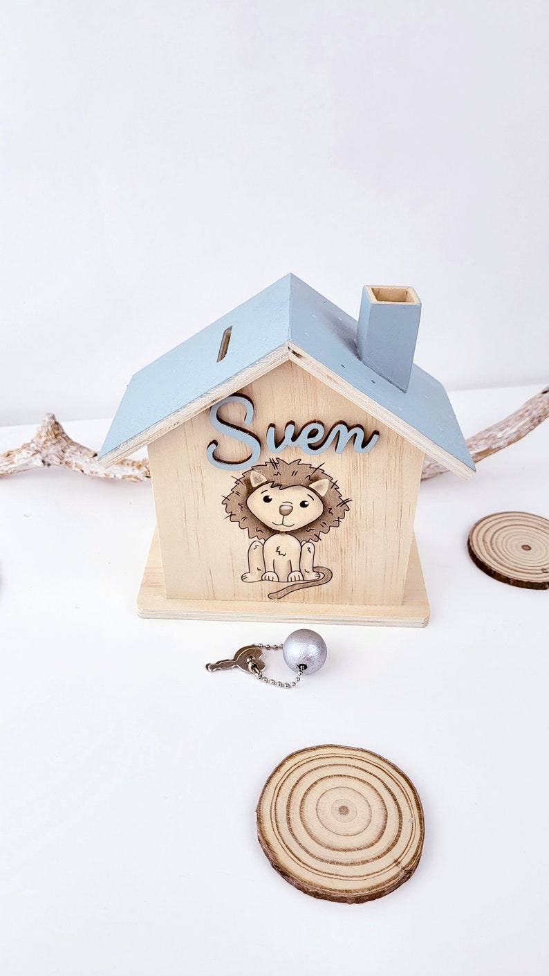 Peut inclure: Une tirelire en bois en forme de maison avec un toit bleu clair et une chemin&eacute;e. Le devant de la tirelire a une illustration de lion et le nom "Sven" &eacute;crit dessus. La tirelire est livr&eacute;e avec une cl&eacute; et une boule argent&eacute;e sur une cha&icirc;ne.