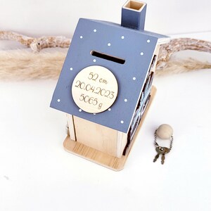 May include: A blue wooden house-shaped piggy bank with white polka dots. The piggy bank has a slot on the roof and a round wooden plaque on the front that says "52 cm 20.04.2023 5065 g". The piggy bank comes with two keys and a wooden bead on a chain.