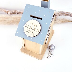 Peut inclure: Une tirelire en bois en forme de maison de couleur bleu clair avec un motif de pois blancs. La tirelire a une fente sur le toit et une petite chemin&eacute;e. Une plaque ronde en bois avec le texte "52 cm 20.04.2023 5065 g" est fix&eacute;e &agrave; l'avant de la tirelire. Une cl&eacute; argent&eacute;e et une boule argent&eacute;e sont &agrave; c&ocirc;t&eacute; de la tirelire.