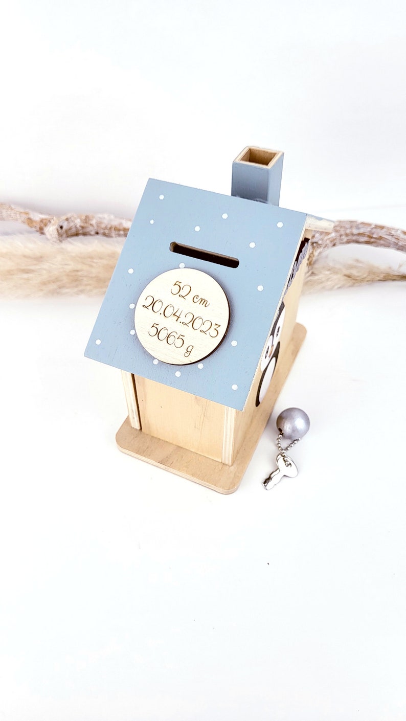 Peut inclure: Une tirelire en bois en forme de maison de couleur bleu clair avec un motif de pois blancs. La tirelire a une fente sur le toit et une petite chemin&eacute;e. Une plaque ronde en bois avec le texte "52 cm 20.04.2023 5065 g" est fix&eacute;e &agrave; l'avant de la tirelire. Une cl&eacute; argent&eacute;e et une boule argent&eacute;e sont &agrave; c&ocirc;t&eacute; de la tirelire.