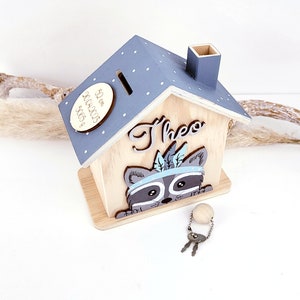 May include: A wooden house-shaped piggy bank with a raccoon face on the front. The house has a blue roof with white polka dots and a small chimney. The raccoon is wearing a blue headband with feathers. The piggy bank has a slot on the roof for coins and comes with a key. The piggy bank is labeled "Theo" and "52 cm 3000.0003 360g".