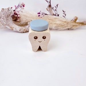 May include: A wooden tooth-shaped box with a light blue lid. The tooth has a smiling face with two eyes. The box is sitting on a white surface.