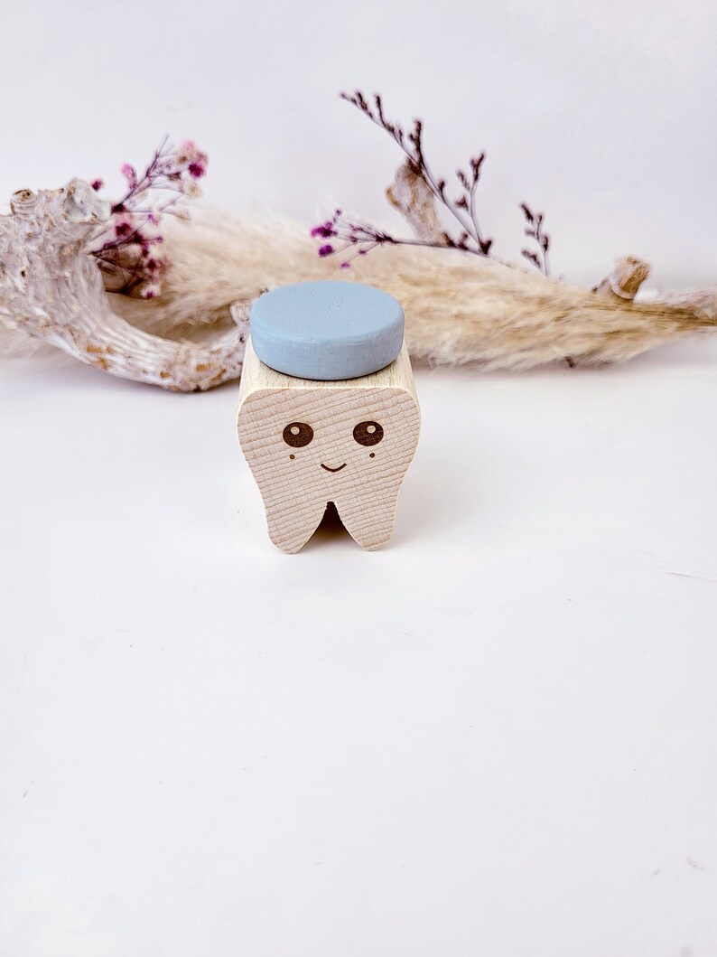 May include: A wooden tooth-shaped box with a light blue lid. The tooth has a smiling face with two eyes. The box is sitting on a white surface.