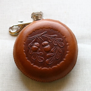 Puede incluir: Una petaca de cuero marrón con una tapa plateada y una cadena. La petaca tiene un diseño en relieve de bellotas y hojas de roble.