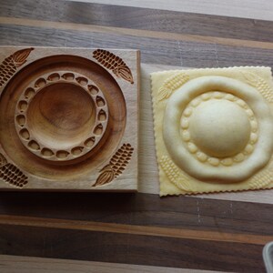 Könnte beinhalten: Ein quadratischer Ravioli-Stempel aus Holz mit einer geschnitzten kreisförmigen Mitte und vier Maiskolben-Motiven an den Ecken, neben einem Stück rohem Nudelteig, der mit demselben kreisförmigen und Maiskolben-Muster geprägt wurde.