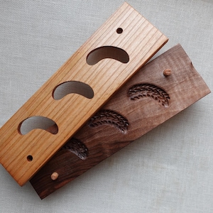 May include: Two wooden wall-mounted key holders. One is light-colored with three curved cutouts. The other is dark brown with three carved wheat stalk designs. Both have small pegs.