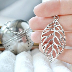May include: Silver tea infuser in the shape of a leaf with a chain attached. The infuser is being held in a hand.