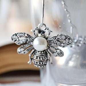 May include: Silver bee charm with a white pearl in the center and clear rhinestones on the wings. The charm is hanging from a silver chain.