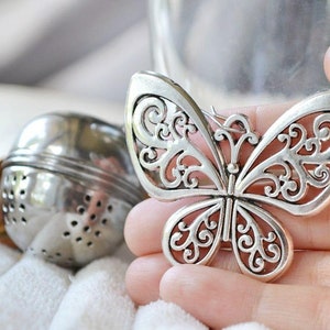 May include: Silver butterfly tea infuser with intricate filigree details. The infuser is held in a hand, ready to be placed in a teacup.