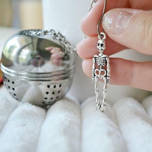 Op de afbeelding: Een zilveren thee-ei in de vorm van een bol met een skeletbedel aan de ketting. De thee-ei heeft een gaasbodem en een deksel.