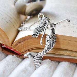 Peut inclure: Infuseur à thé en forme de sirène argentée avec une chaîne attachée à une boule à thé en argent. L'infuseur est posé sur un livre ouvert.