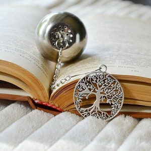 May include: Silver tea infuser with a chain and a tree of life design. The infuser is resting on an open book.