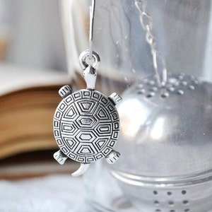 May include: A silver-colored tea infuser in the shape of a turtle. The turtle has a detailed shell pattern and is suspended by a chain. The infuser is next to a stainless steel tea ball.