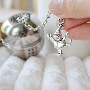 以下が含まれることがあります： 雪だるまのチャームが付いたシルバーのティーインフューザー。雪だるまは帽子とマフラーを着用し、両手に棒を持っています。