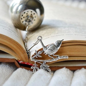 May include: A silver tea infuser shaped like a bird on a branch with leaves. The infuser is hanging from an open book.