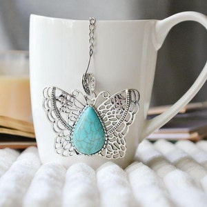 May include: A silver butterfly tea infuser with a turquoise gemstone pendant. The infuser is hanging from a white ceramic mug.