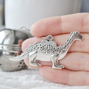 May include: A silver-colored dinosaur pendant with a detailed design. The pendant features a long neck, a spiky back, and a small loop for attaching to a chain or other jewelry. The dinosaur is held in a hand.