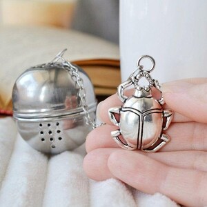 May include: Silver tea infuser with a beetle shaped handle. The infuser is round and has a mesh bottom. The beetle is detailed and has a shiny finish.