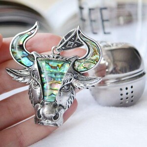 May include: A silver-toned tea infuser in the shape of a bull's head, featuring iridescent abalone shell accents. The bull's head is detailed with horns and facial features. A separate, spherical tea infuser is in the background.