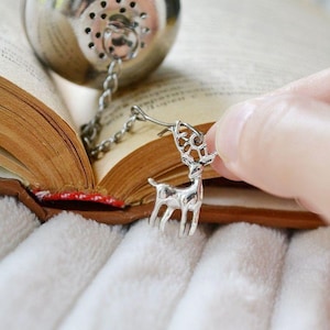 May include: A silver tea infuser with a chain attached to a silver reindeer charm. The infuser is sitting on an open book.